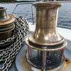 Bronze winch on a boat with chain, water, and trees in the background