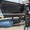 Man washing a black sailboat with a bottle of auto shampoo on a dock.