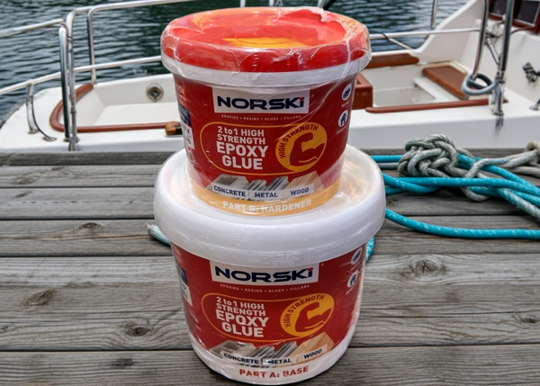 Two containers of Norski epoxy glue on a wooden dock with water and a boat in the background.