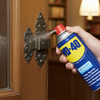 Hand using a can of WD-40 on a wooden door hinge with a bookshelf background
