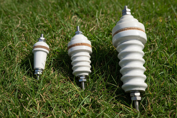 Three ceramic high-voltage bushings in different sizes arranged on a plain light background, featuring a ribbed white body, a brown insulating ring, and silver metal terminal hardware.