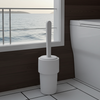 White toilet brush holder with a toilet brush on a tiled bathroom floor, window in the background showing water view.