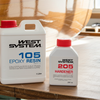 West System epoxy resin and hardener bottles on a wooden surface with a person working in the background.