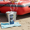 Person cleaning an inflatable boat on a beach with a bottle of cleaner and a bucket nearby.