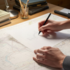 Person working on a map with a pen at a desk under a lamp